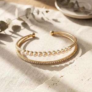 Könnte beinhalten: Ein goldfarbenes, offenes Armband mit zwei getrennten Reifen. Ein Reifen ist mit einer Reihe kleiner, runder, weißer Perlen besetzt, während der andere Reifen mit einer durchgehenden Linie aus klaren, funkelnden Kristallen verziert ist. Das Armband hat abgerundete Enden.