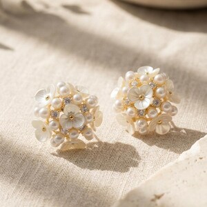 May include: A pair of gold-tone stud earrings featuring a cluster design. Each earring is composed of small white floral-shaped components, round white pearls, and tiny clear sparkling crystals arranged in a spherical, bouquet-like formation.