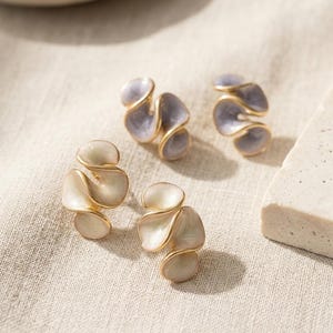 May include: Close-up of two pairs of earrings. One pair is a light lavender color, and the other is a creamy white. Both pairs have a gold-colored metal trim. The earrings have a unique, layered petal design. The earrings are on a neutral-colored fabric.