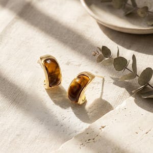 Könnte beinhalten: Ein Paar goldfarbene Creolen mit braunem, marmoriertem Steineinsatz. Die Ohrringe liegen auf einer weißen Oberfläche mit Schatten und einem Zweig Grünzeug im Hintergrund. Die Ohrringe haben ein geschwungenes Design.
