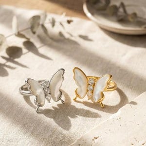 May include: Two butterfly rings, one silver and one gold, are displayed on a neutral surface. The butterfly wings are white, and the rings feature clear gemstones. The gold ring has a warm, shiny finish.