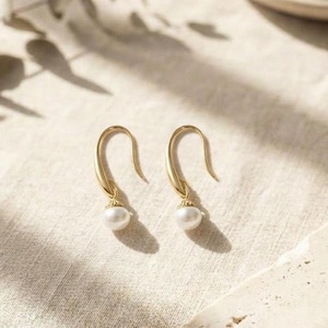 May include: A pair of gold-colored hook earrings with a small, round, white pearl dangling from each. The earrings are displayed on a light-colored, textured surface, with soft shadows.