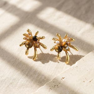 May include: A pair of gold-toned earrings with a unique design. Each earring features a cluster of brown, black, and clear beads, along with a small white pearl. The earrings have a curved, open design.