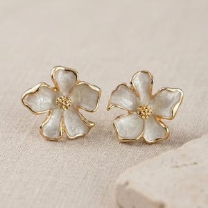 以下が含まれることがあります： 花型のスタッドピアス。各イヤリングは、金色のトリムが付いた白いエナメルペタルデザインが特徴です。各花の中心は、テクスチャ加工された金色のビーズです。イヤリングはニュートラルなトーンの表面に表示されます。
