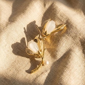 May include: A gold-toned floral brooch featuring two white flower buds with crystal accents and a pearl. The brooch is designed in a botanical style, with delicate leaves and stems. The jewelry is set against a neutral fabric background.
