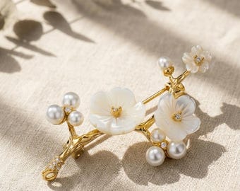 May include: A gold-toned brooch shaped like a flowering branch, featuring two white mother-of-pearl flowers with small clear crystals at their centers, several round white pearls, and additional small clear crystals embedded along the stem.