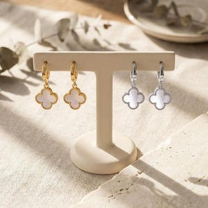 Op de afbeelding: Twee paar klavervormige oorbellen op een displaystandaard. Eén paar is goudkleurig met een witte kern, de andere is zilverkleurig met een witte kern. De oorbellen staan op een beige standaard, met een neutrale achtergrond.