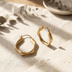 May include: A pair of gold-toned hoop earrings featuring an irregular, organic, and wavy shape. The earrings have a matte finish and a hinged post closure mechanism, designed to be worn through the earlobe with a secure and functional clasp.