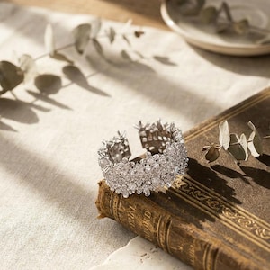 May include: A silver-colored cuff bracelet with a floral design, resting on a vintage book. The bracelet is encrusted with small, sparkling stones, and the book has gold lettering. The background includes a neutral-toned cloth and a small plate.