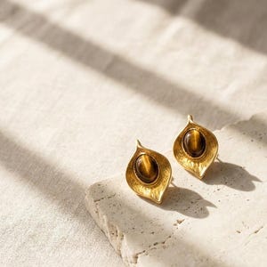 May include: A pair of gold-colored stud earrings with a leaf-like design. Each earring features a brown, oval-shaped stone set in the center. The earrings are displayed on a textured, off-white surface, with soft shadows.