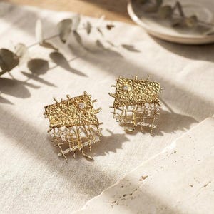 May include: A pair of gold-toned earrings with a textured, rectangular top section and a delicate, grid-like design below. Small, clear gemstones are set within the grid. The earrings are displayed on a neutral-toned surface.