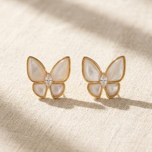 May include: A pair of butterfly-shaped earrings. Each earring features a gold-colored frame with white, pearlescent wings and a clear, oval-shaped gem at the center. The earrings are displayed on a neutral-colored, textured surface.