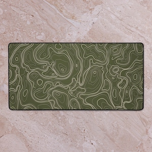 May include: A rectangular desk mat featuring a topographic map design. The mat has a dark olive green background with a pattern of beige contour lines. The mat has a black border and is designed for use on a desk.