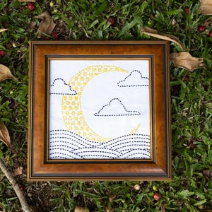 May include: A framed artwork featuring a crescent moon and clouds. The moon is yellow with a star pattern, and the clouds and waves are outlined in black stitching. The frame is brown.