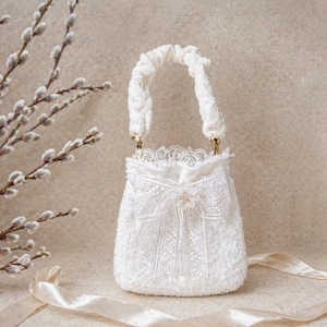 Könnte beinhalten: Eine weiße, strukturierte Handtasche mit geflochtenem Griff und zarten Spitzendetails. Die Tasche hat eine Schleife und einen dekorativen Verschluss. Im Hintergrund befinden sich Weidenkätzchen und ein Satinband.