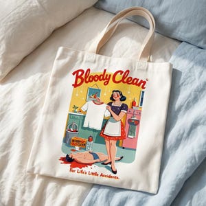 Puede incluir: Bolso tote de lona blanquecina con asas color crema. El bolso presenta una ilustración de estilo retro con el texto "Bloody Clean" en rojo. La ilustración representa a una mujer en una escena de cocina con una persona en el suelo y el texto "For Life's Little Accidents."