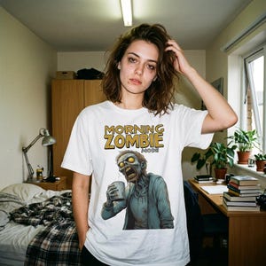 Könnte beinhalten: Weißes T-Shirt mit dem Text "MORNING ZOMBIE MODE" in Gold und einer Cartoon-Zombie-Grafik, die eine Tasse hält. Der Zombie ist in Blau-, Grün- und Grautönen dargestellt. Das T-Shirt wird von einer Person getragen.