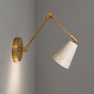 May include: A wall-mounted lamp featuring a brass finish arm with two adjustable joints and a circular wall plate. The lamp has a conical white shade with a series of small circular perforations along the bottom edge, directing light downward.