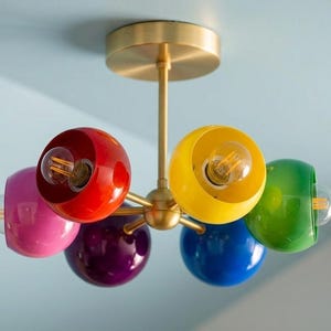 Può includere: Un moderno lampadario a soffitto con sei paralumi sferici colorati in rosso, rosa, viola, blu, giallo e verde. L'apparecchio ha una finitura in ottone e lampadine a vista.