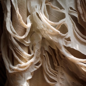 May include: Close-up of a cream-colored fabric with a distressed design. The fabric has multiple cut-out sections, creating an open, textured pattern. The background is a neutral beige, highlighting the intricate details of the fabric.