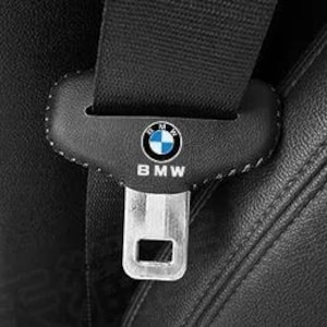 Peut inclure: Une housse de ceinture de sécurité en cuir noir arborant le logo et le texte BMW en blanc. La housse est cousue avec un fil blanc et est placée sur une sangle de ceinture noire, avec une boucle métallique argentée visible en bas.