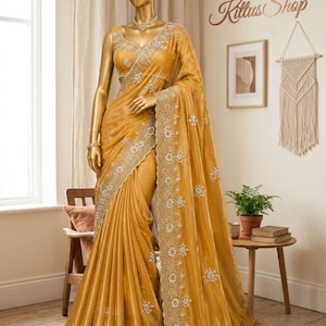 May include: A mustard yellow saree draped on a gold mannequin, featuring intricate silver embroidery along the borders and scattered floral motifs throughout the fabric. The garment includes a matching sleeveless blouse with a V-neckline and silver detailing.