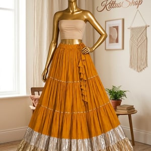 Puede incluir: Una falda larga de niveles color amarillo mostaza con diseño de cintura alta y detalle de lazo lateral, acentos horizontales plateados entre niveles y un dobladillo ancho con volantes en plata y oro, junto a un top corto beige sin tirantes.