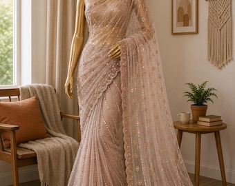 May include: A blush-colored saree with intricate gold sequin detailing is draped on a mannequin. The sheer fabric features a delicate floral pattern and a scalloped hem. The saree is displayed in a well-lit room with a neutral color palette.