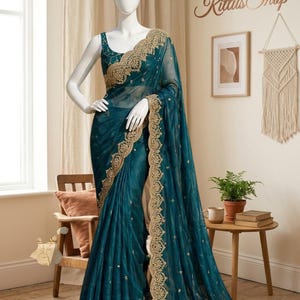 May include: A teal saree with gold embroidery is displayed on a mannequin. The saree features intricate gold detailing along the borders and bodice. The mannequin is in a room with neutral-toned walls and decor, including a small table with books and a plant.