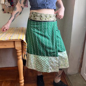 May include: A knee-length wrap skirt featuring a green fabric with white vertical floral stripes, a wide waistband with a light green patterned fabric and silver metal studs, and a black horizontal zipper detail across the upper section.