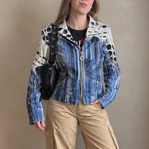 Puede incluir: Una chaqueta de manga larga con un patrón de rayas verticales azules y blancas en el torso y las mangas, con hombros blancos adornados con puntos negros circulares y alargados. La chaqueta tiene cierre de cremallera y se combina con pantalones cargo beige.