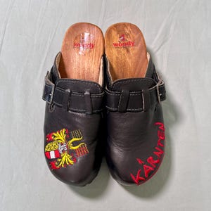 May include: A pair of black leather clogs with wooden soles. The clogs feature a buckle strap and embroidered details, including a coat of arms and the word "Kärnten" in red. The insoles are branded with the word "woody".