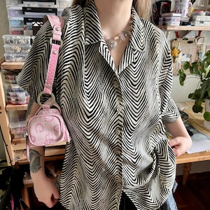 May include: A black and white patterned shirt with a zig-zag design. The shirt has short sleeves and is buttoned up. A small pink purse with a matching strap is visible.