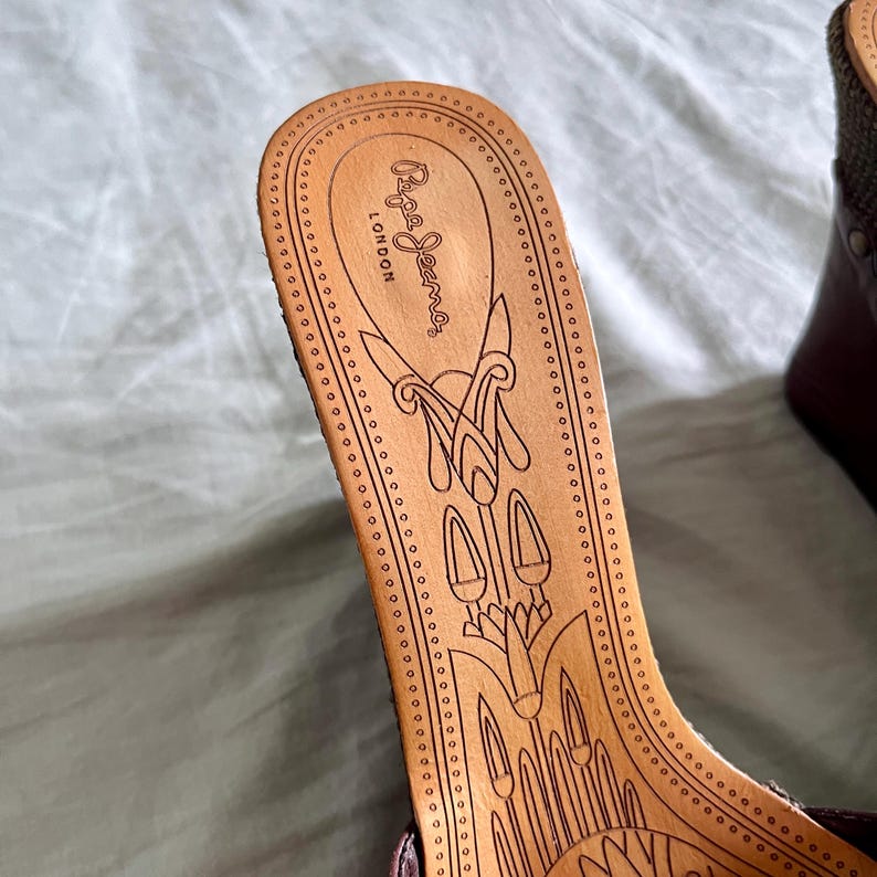 May include: Close-up of a brown wedge sandal's insole. The wooden insole features an engraved design and the brand name "Pepe Jeans London". The insole is surrounded by a line of small, evenly spaced holes.