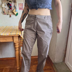 Op de afbeelding: Licht taupekleurige broek met een ritssluiting aan de voorzijde, een tailleband met knoop en zijzakken. De broek heeft een recht model en een klein, rond logopatch in het zwart geborduurd op het bovenste gedeelte van de linker dij.