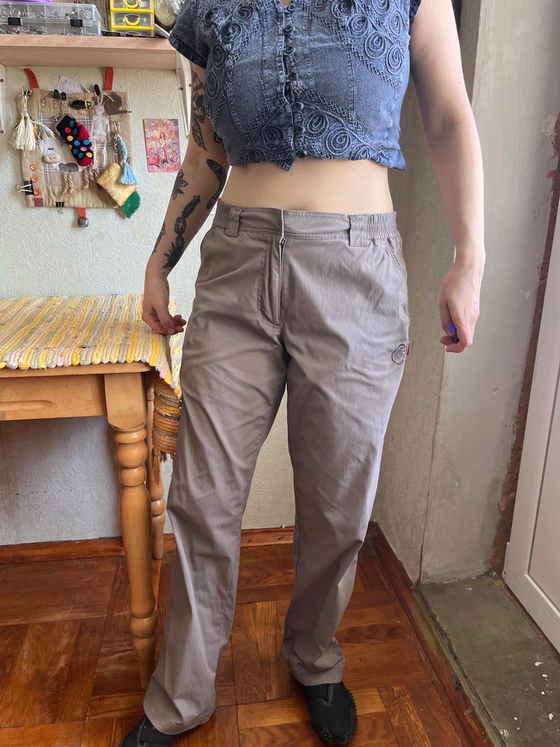 Op de afbeelding: Licht taupekleurige broek met een ritssluiting aan de voorzijde, een tailleband met knoop en zijzakken. De broek heeft een recht model en een klein, rond logopatch in het zwart geborduurd op het bovenste gedeelte van de linker dij.