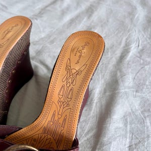 May include: Close-up of two maroon wedge sandals with woven fabric and gold-colored studs. The footbed is tan leather with a decorative design and the brand name "Pepe Jeans London".