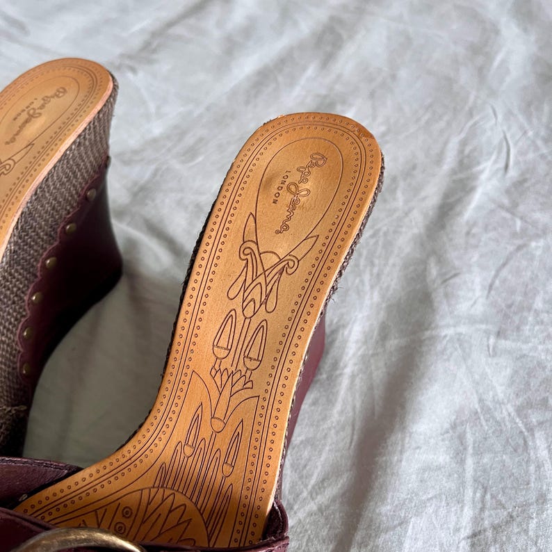 May include: Close-up of two maroon wedge sandals with woven fabric and gold-colored studs. The footbed is tan leather with a decorative design and the brand name "Pepe Jeans London".