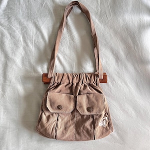May include: A tan suede shoulder bag with a wooden handle and two front pockets. The bag has a drawstring closure and a small embroidered logo on the front. The bag's strap is made of the same suede material.