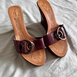 May include: A pair of burgundy wedge sandals with a leather strap and a large gold-colored buckle. The footbed is tan with a decorative pattern and the brand name "Pepe Jeans London" is visible.