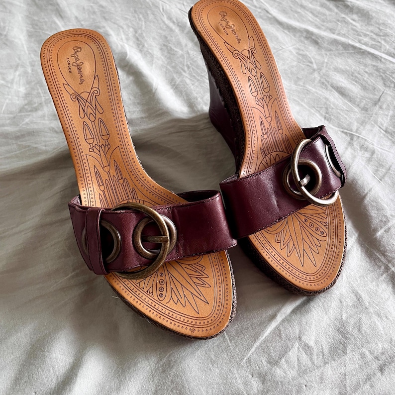 May include: A pair of burgundy wedge sandals with a leather strap and a large gold-colored buckle. The footbed is tan with a decorative pattern and the brand name "Pepe Jeans London" is visible.