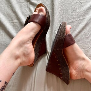 May include: A pair of brown wedge sandals with leather straps and a woven texture on the soles. The sandals have a buckle detail and are set against a neutral background.