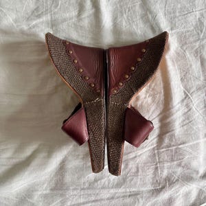 May include: A pair of burgundy and brown wedge sandals. The sandals feature a woven fabric design with leather accents and small gold-colored studs. The open-toe shoes have a wedge heel and a strap that buckles around the ankle.