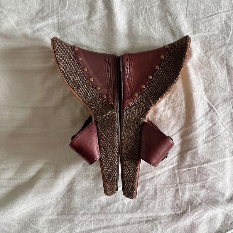 May include: A pair of burgundy and brown wedge sandals. The sandals feature a woven fabric design with leather accents and small gold-colored studs. The open-toe shoes have a wedge heel and a strap that buckles around the ankle.