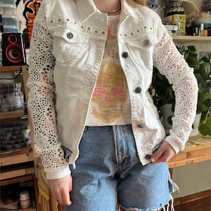 Puede incluir: Chaqueta vaquera blanca con mangas de encaje y botones decorativos. La chaqueta se combina con una camiseta gráfica y pantalones cortos de mezclilla. El estilo general es casual y de inspiración vintage.