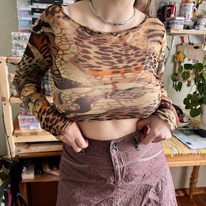 May include: A long-sleeved, sheer top with a brown and orange animal print pattern. The top is paired with a maroon skirt with a subtle plaid design. A silver necklace is also visible.