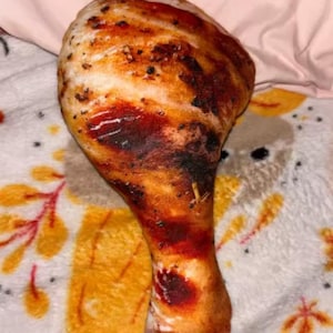 May include: A plush, oversized chicken leg pillow. The pillow features a realistic design with brown, red, and white coloring to mimic a roasted chicken leg. The pillow is resting on a patterned blanket with a pink background.