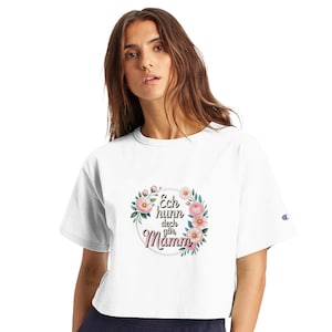 Può includere: Top corto bianco con un design floreale e la scritta "Ech hunn dech gär Mamm" in una cornice circolare. Il design include fiori rosa e foglie verdi. La maglietta ha un logo Champion sulla manica.