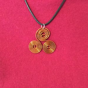 Peut inclure: Un collier avec pendentif de couleur cuivre, orné d'un motif à triple spirale. Le pendentif est suspendu à un cordon noir. Le collier est présenté sur un fond rouge.