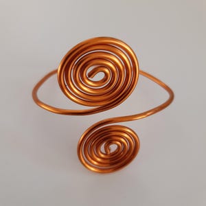Puede incluir: Brazalete de color cobre con dos diseños en espiral. El brazalete está hecho de una sola pieza de alambre, con una espiral grande en la parte superior y otra más pequeña en la parte inferior. El diseño es simple y elegante.